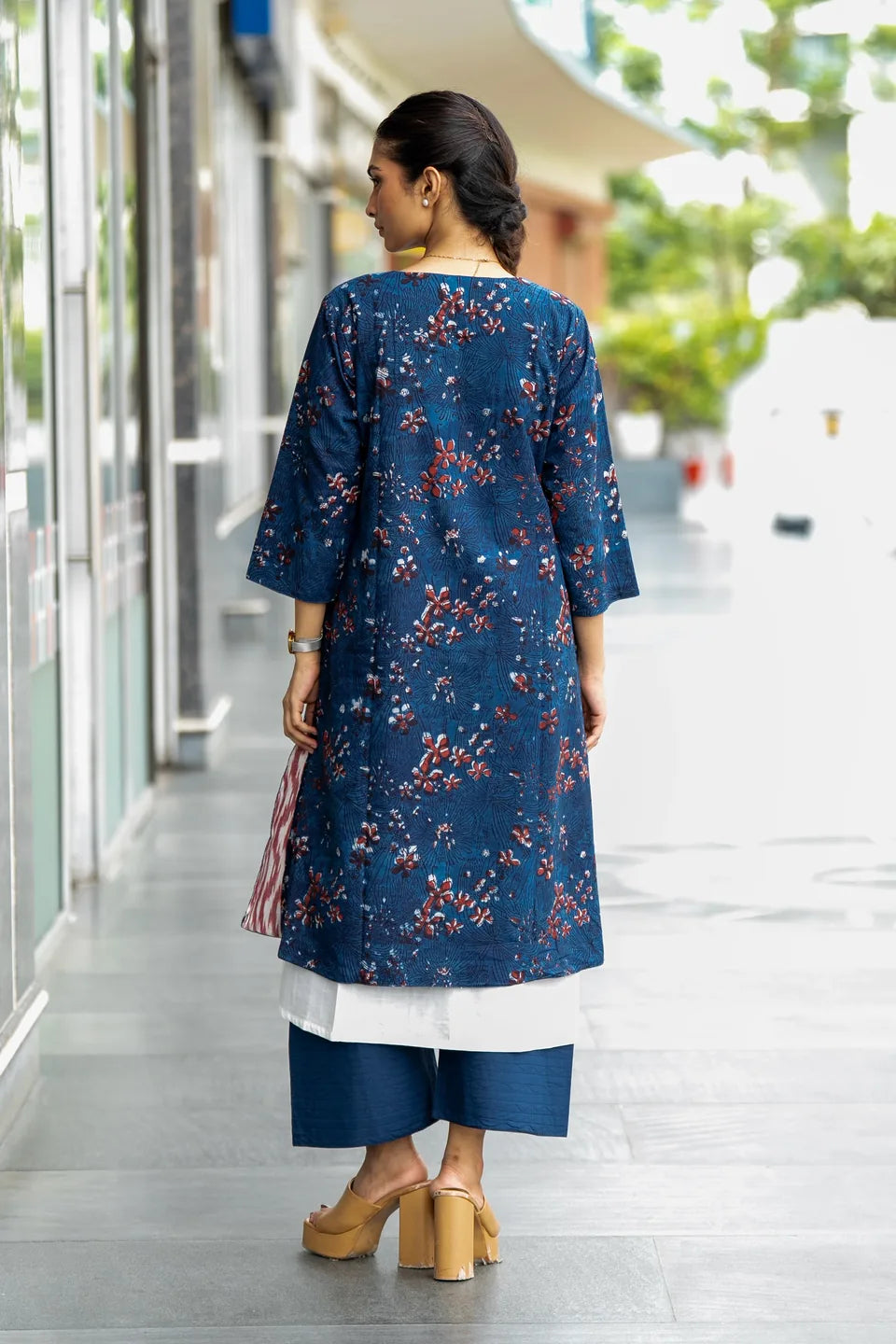 A woman viewed from behind wearing a navy blue floral printed kurta with three-quarter sleeves, paired with a white underskirt and navy cropped pants, standing in a modern outdoor walkway.